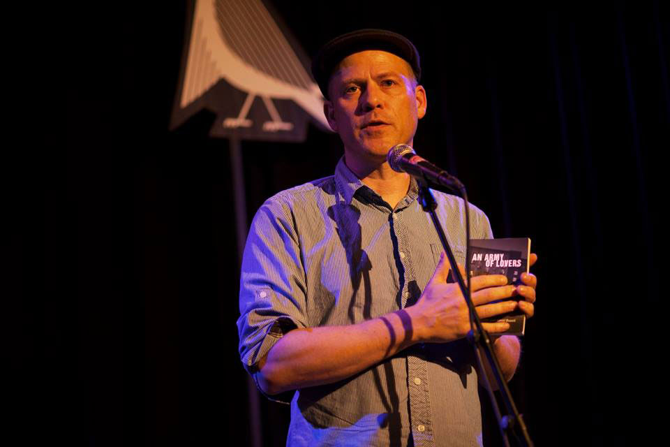 Photo of a man at a microphone. He is holding a book, An Army of Lovers.