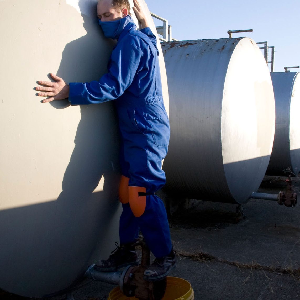 A man in a blue jumpsuit with kneepads scaling industrial tanks.